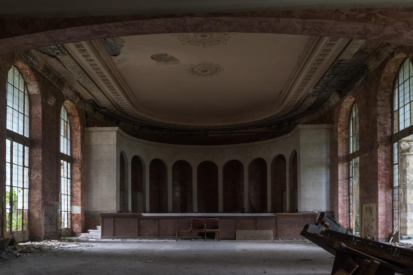 El gran salón abandonado del Sanatorio Metallurg en Tskaltubo, con altos ventanales en arco, paredes de mármol y un escenario curvo